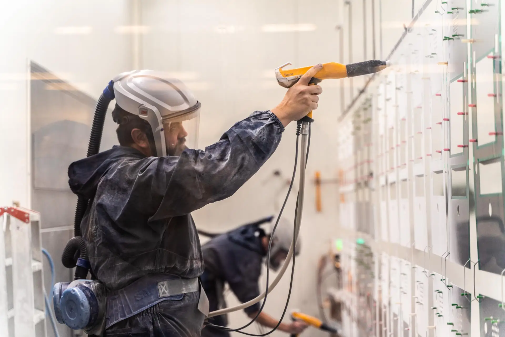Mitarbeiter im Schutzanzug lackiert Metallplatten mit einer Pulverbeschichtungspistole in einer Lackierkabine.