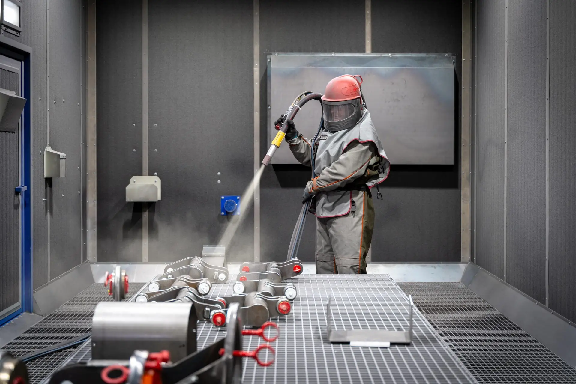 Person in Schutzkleidung sandstrahlt Metallteile in einer geschlossenen Kabine.