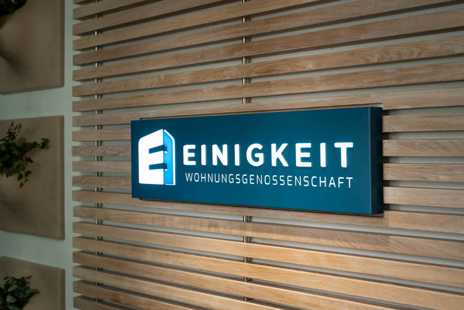 Leuchtendes Firmenschild mit dem Logo der Einigkeit Wohnungsgenossenschaft an einer Wand aus Holzlamellen.
