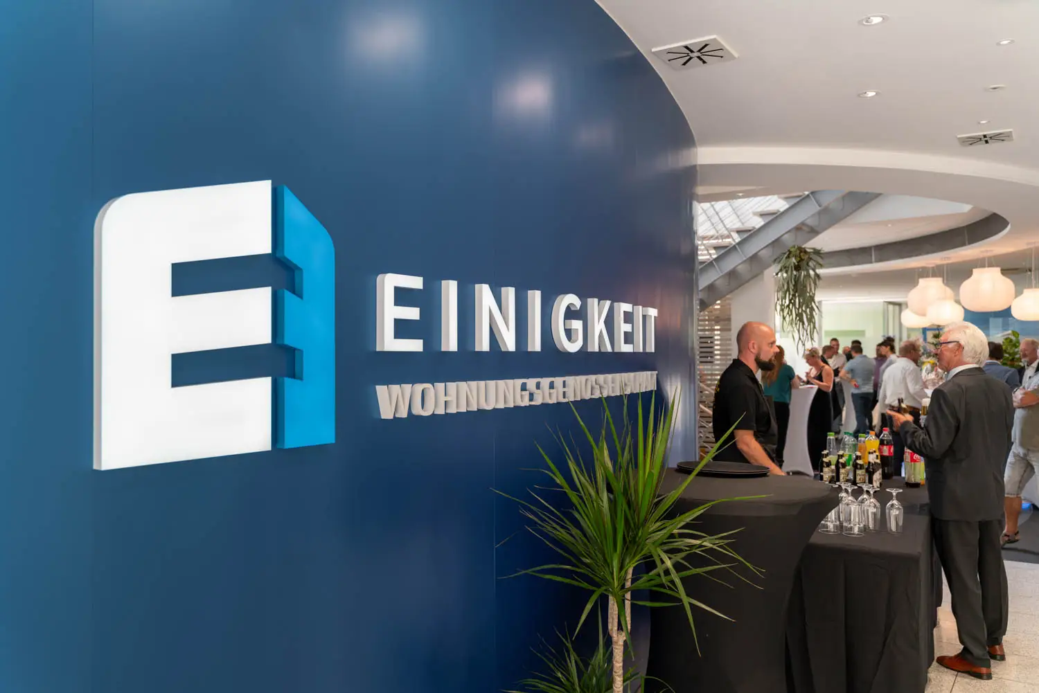 mpfangsbereich der Einigkeit Wohnungsgenossenschaft mit blauem Logo an der Wand und Gästen bei einer Veranstaltung.
