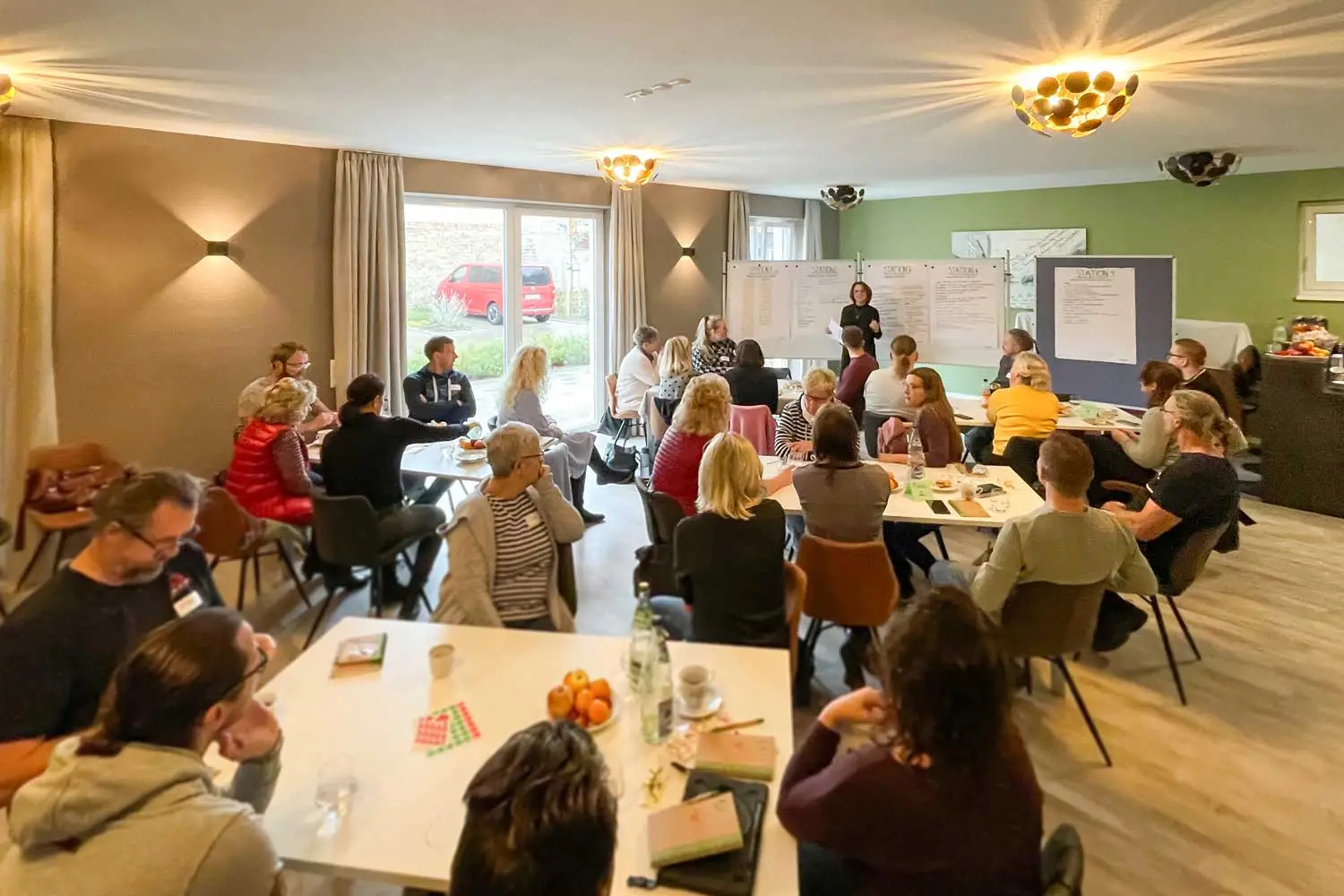 Gruppenraum mit Teilnehmenden, die einer Referentin zuhören, während sie Ergebnisse an Pinnwänden präsentiert.