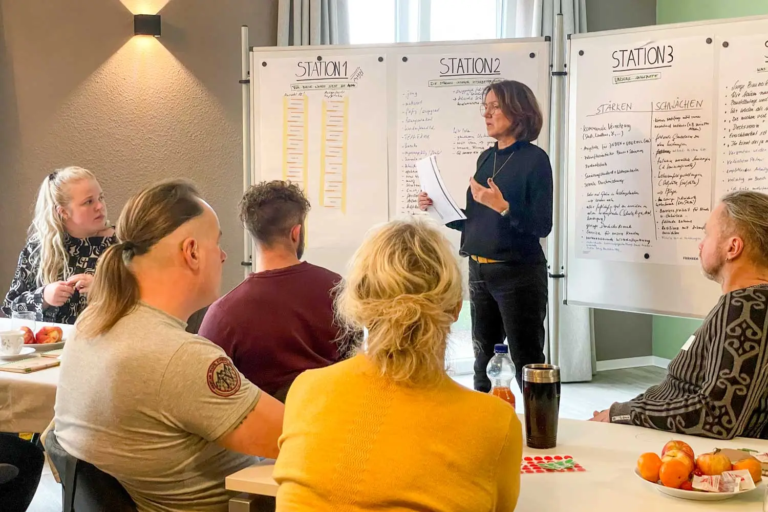 Frau hält eine Präsentation vor Workshop-Teilnehmenden, die an Tischen sitzen und Notizen machen.