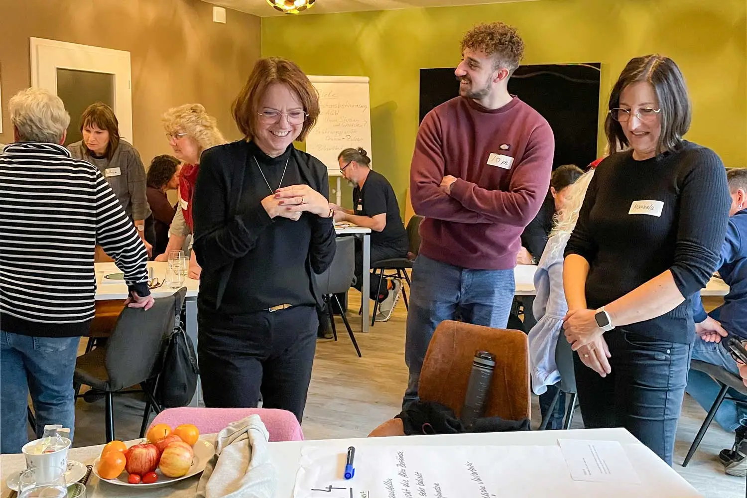 Drei Personen unterhalten sich lachend während eines Workshops, im Hintergrund arbeiten weitere Teilnehmende in Gruppen.
