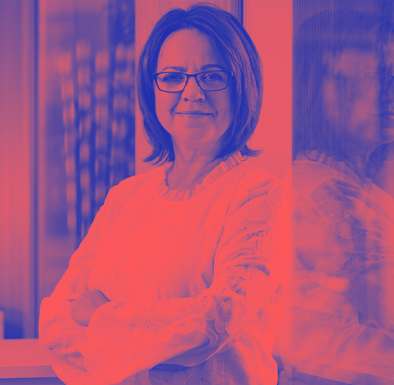 Iris Palme mit heller Bluse steht mit verschränkten Armen an einem Fenster und blickt freundlich in die Kamera.