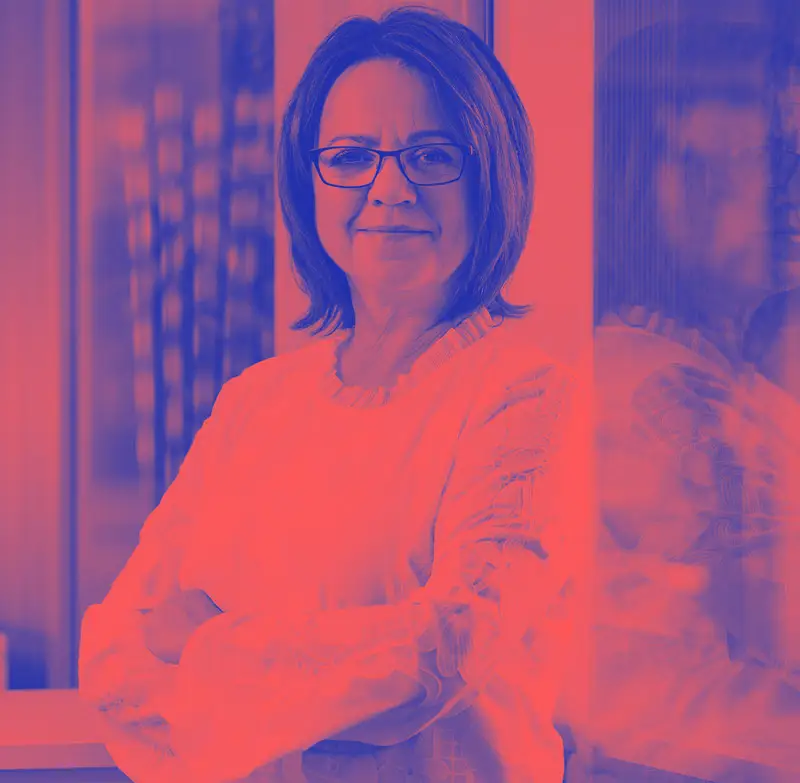 Iris Palme mit heller Bluse steht mit verschränkten Armen an einem Fenster und blickt freundlich in die Kamera.